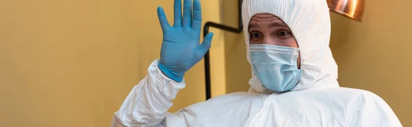 Panoramic crop of man in medical mask, hazmat suit and latex glove waving hand at home — Stock Photo