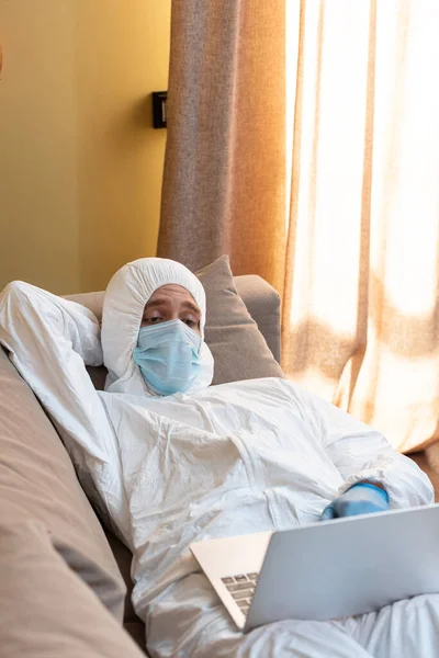 Foco seletivo de freelancer em máscara médica e terno hazmat usando laptop enquanto deitado no sofá em casa — Fotografia de Stock