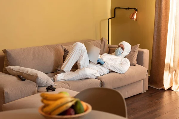 KYIV, UKRAINE - APRIL 24, 2020: Selective focus of man in hazmat suit and medical mask using laptop near joystick and remote controller on couch — Stock Photo