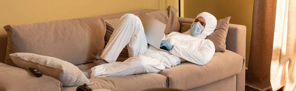 KYIV, UKRAINE - APRIL 24, 2020: Panoramic shot of man in hazmat suit and medical mask using laptop near remote controller and joystick on sofa — Stock Photo