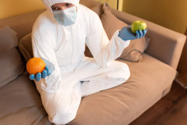 Selective focus of man in hazmat suit, medical mask and latex gloves holding orange and apple on sofa at home — Stock Photo