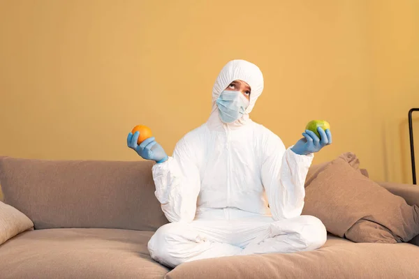 Pensive man in medical mask and hazmat suit holding apple and orange on couch — Stock Photo