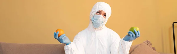 Orientamento panoramico dell'uomo premuroso in tuta hazmat e maschera medica che tiene mela e arancia a casa — Foto stock