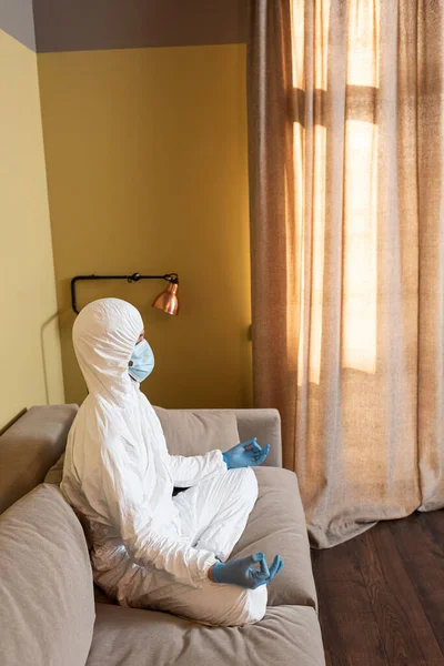 Vista lateral del hombre en traje de materiales peligrosos, guantes de látex y máscara médica meditando en el sofá - foto de stock