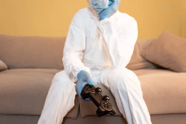 KYIV, UKRAINE - 24 AVRIL 2020 : Mise au point sélective de l'homme en masque médical, gants en latex et combinaison Hazmat tenant un joystick sur le canapé — Photo de stock