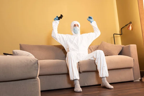 KYIV, UKRAINE - APRIL 24, 2020: Selective focus of excited man in hazmat suit holding joystick on couch — Stock Photo