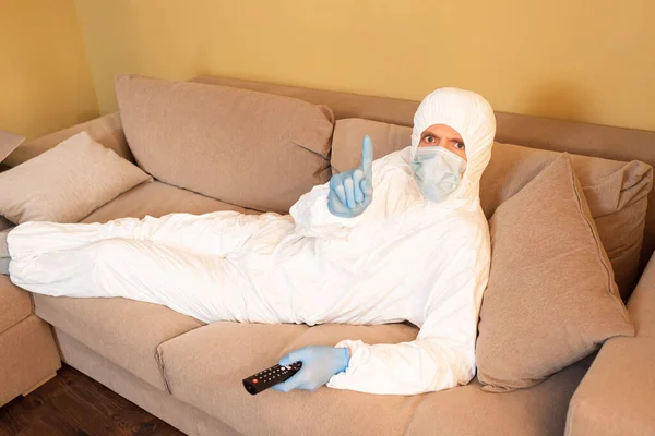 Hombre con traje de materiales peligrosos y máscara médica apuntando con el dedo y sosteniendo el mando a distancia en el sofá - foto de stock