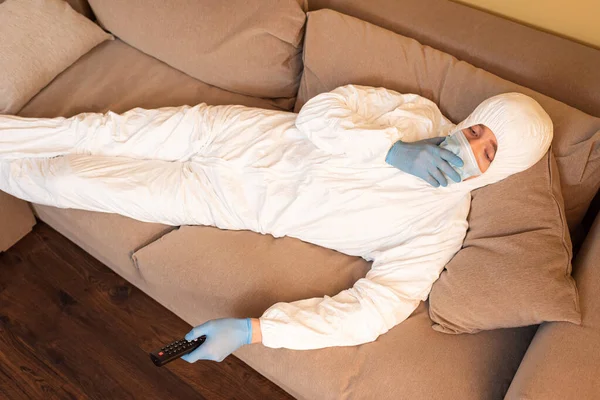 High angle view of man in hazmat suit, medical mask and latex gloves watching film on couch — Stock Photo