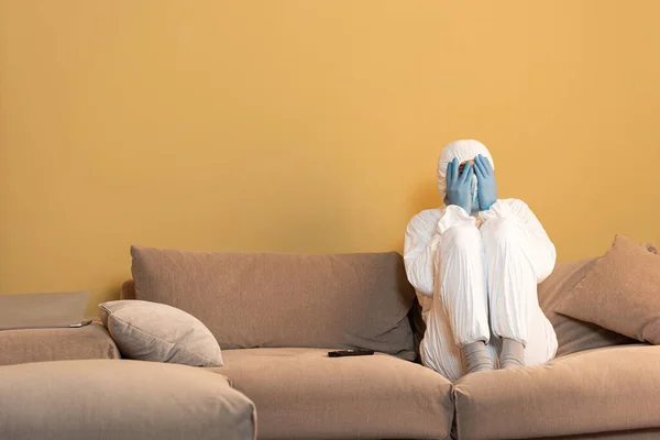 Man in hazmat suit and latex gloves covering face near remote controller on couch — Stock Photo