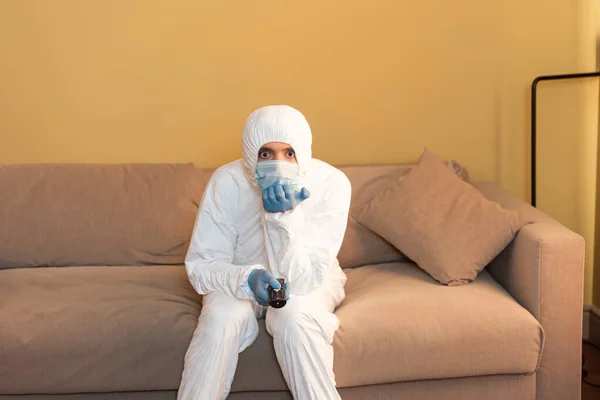 Человек в защитном костюме и медицинская маска щелкают каналами на диване — стоковое фото