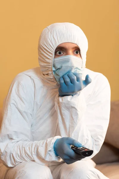 Hombre con traje de materiales peligrosos, máscara médica y guantes de látex que sostiene el mando a distancia en el sofá - foto de stock