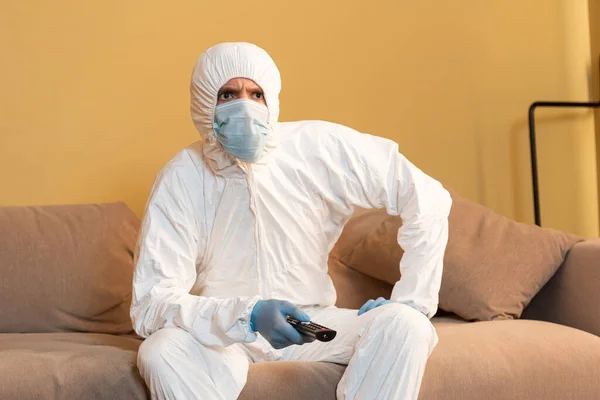 Nachdenklicher Mann in Schutzanzug und medizinischer Maske vor dem Fernseher auf dem Sofa — Stockfoto