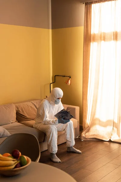 Foco seletivo do homem em terno hazmat e revista de leitura de máscara médica no sofá — Fotografia de Stock