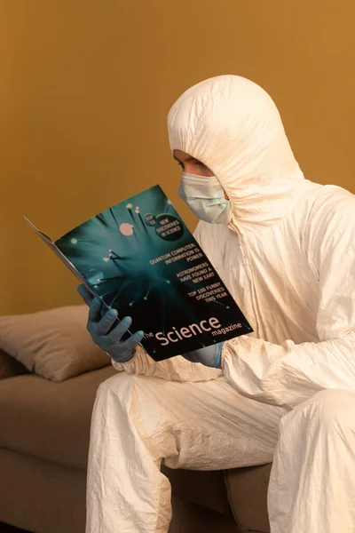 Uomo in maschera medica e tuta hazmat lettura rivista scientifica sul divano a casa — Foto stock