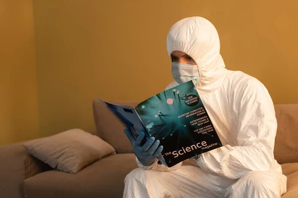 Hombre con traje de materiales peligrosos, máscara médica y guantes de látex leyendo la revista científica en el sofá - foto de stock