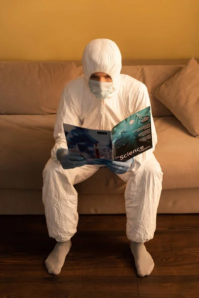 Mann in Schutzanzug und medizinischer Maske liest Wissenschaftsmagazin, während er zu Hause auf der Couch sitzt — Stockfoto