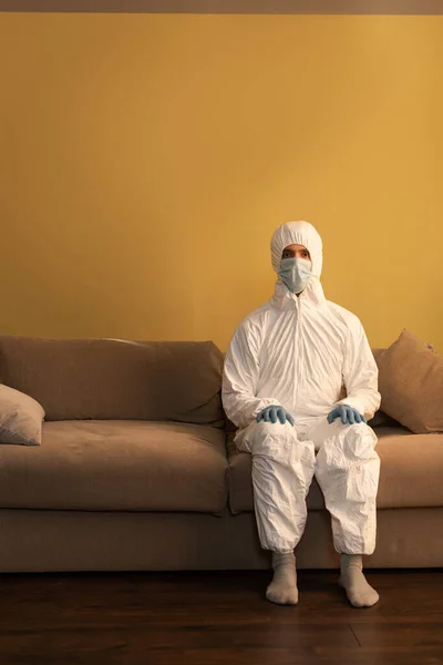 Hombre con traje de materiales peligrosos, máscara médica y guantes de látex mirando a la cámara mientras está sentado en el sofá - foto de stock
