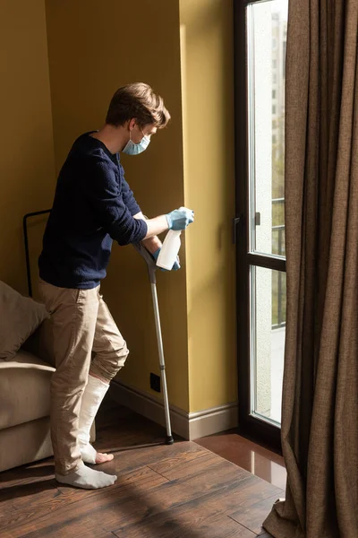 Vista lateral do homem com deficiência em máscara médica e luvas de látex segurando detergente e muleta na sala de estar — Fotografia de Stock