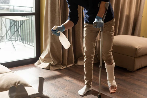 Cropped view of man with broken leg using detergent while holding crutch at home — Stock Photo