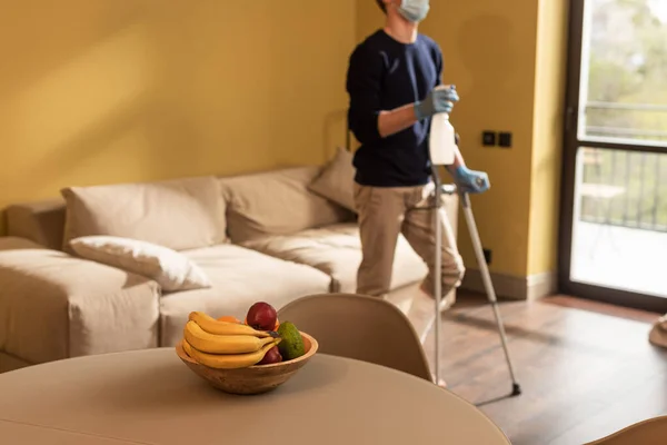 Selektiver Fokus eines behinderten Mannes in medizinischer Maske, der eine Flasche Waschmittel und Krücken im Wohnzimmer hält — Stockfoto