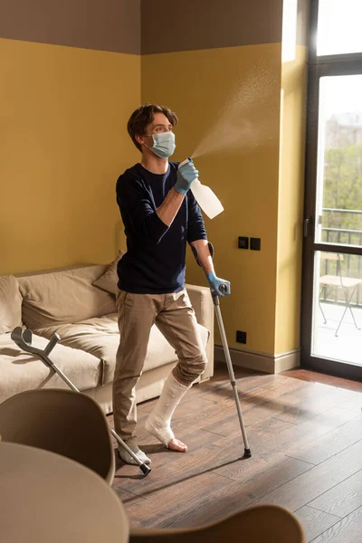 Enfoque selectivo del hombre discapacitado en máscara médica sosteniendo muletas y rociando detergente en casa - foto de stock