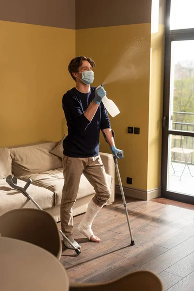 Concentration sélective de l'homme handicapé dans un masque médical tenant des béquilles et pulvérisant du détergent à la maison — Photo de stock