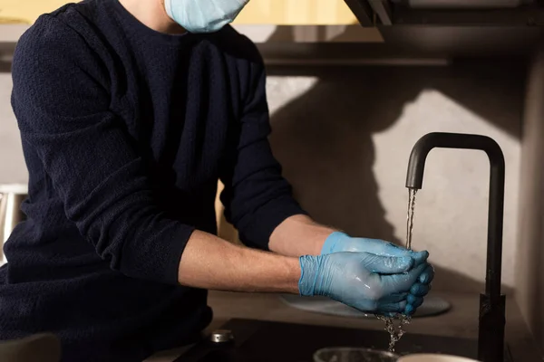 Vista recortada del hombre en máscara médica y guantes de látex lavándose las manos en la cocina - foto de stock