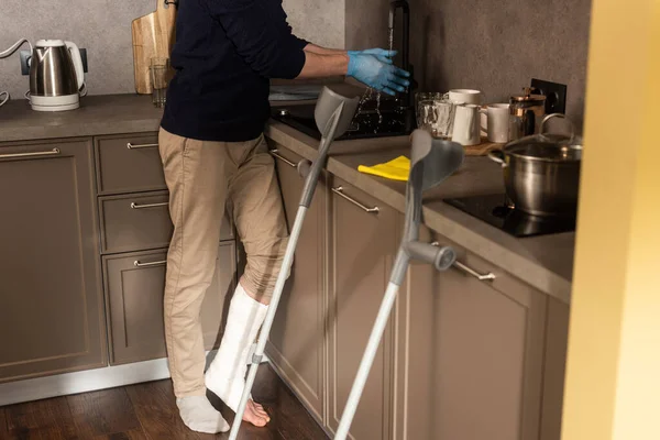 Vista recortada del hombre con vendaje de yeso en la pierna lavándose las manos en guantes de látex en la cocina - foto de stock