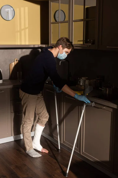 Seitenansicht eines Mannes mit gebrochenem Bein, der Küchenarbeitsplatte in der Nähe von Krücken putzt — Stockfoto