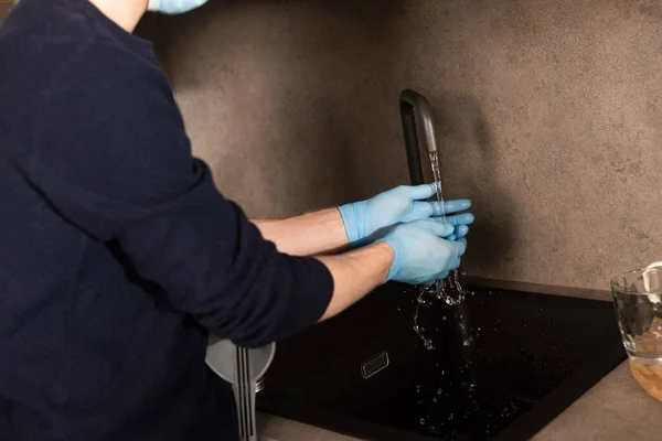 Vista recortada del hombre en guantes de látex y máscara médica lavándose las manos cerca de la muleta en la cocina - foto de stock