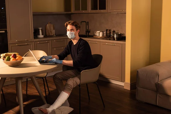 Vista laterale dell'uomo in maschera medica e bendaggio in gesso su led utilizzando laptop in cucina — Foto stock