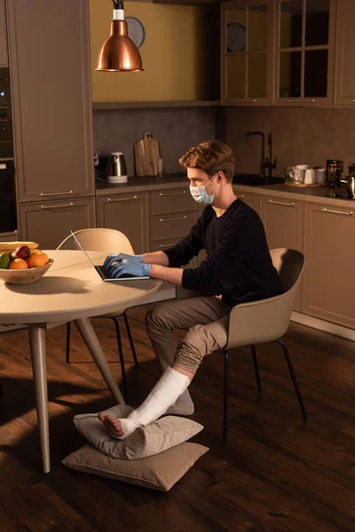 Vista lateral do freelancer em máscara médica com perna quebrada trabalhando no laptop na cozinha — Fotografia de Stock
