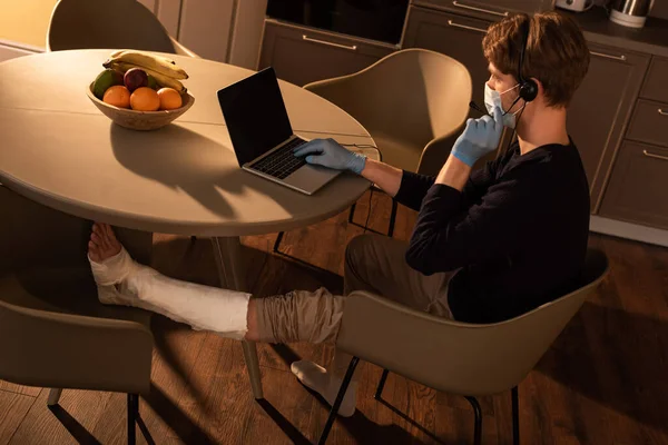 Vista laterale del libero professionista in maschera medica con gamba rotta che lavora con auricolare e laptop sul tavolo da cucina — Foto stock