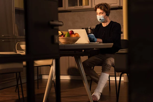 Enfoque selectivo del hombre en máscara médica y vendaje de yeso en la pierna usando auriculares y computadora portátil en la cocina - foto de stock