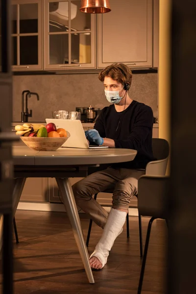 Enfoque selectivo del hombre discapacitado en máscara médica usando auriculares y portátil cerca de frutas en la mesa de la cocina - foto de stock