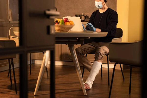 Selective focus of man in medical mask and plaster bandage on leg using headset and laptop in kitchen — Stock Photo