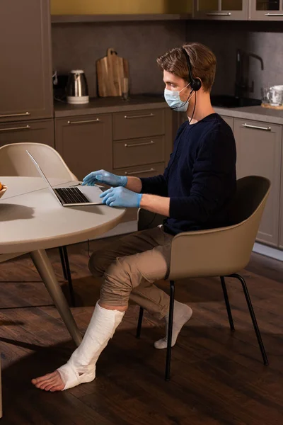 Vue latérale de l'homme handicapé en masque médical et gants en latex travaillant avec ordinateur portable et casque sur la table de cuisine — Photo de stock
