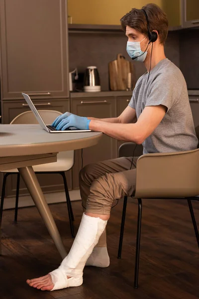 Vue latérale du télétravailleur en masque médical, gants en latex et bandage de plâtre sur la jambe à l'aide d'un casque tout en travaillant sur un ordinateur portable dans la cuisine — Photo de stock