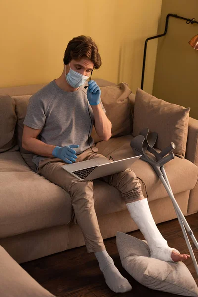 Homem em máscara médica e bandagem de gesso na perna usando fone de ouvido enquanto trabalhava no laptop no sofá — Fotografia de Stock