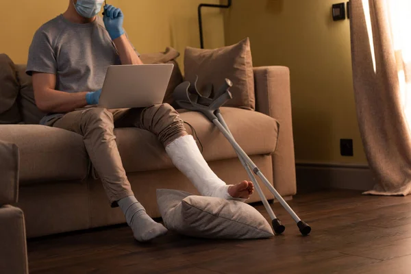Vista cortada do homem com perna quebrada e máscara médica usando laptop e fone de ouvido perto de muletas no sofá — Fotografia de Stock