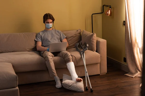 Homem em máscara médica com perna quebrada usando fone de ouvido enquanto trabalhava no laptop em casa — Fotografia de Stock