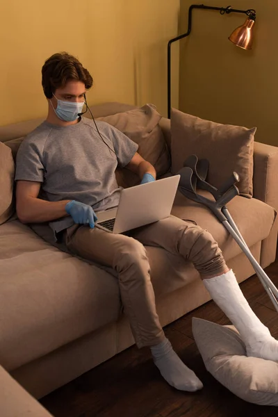 Homme handicapé en masque médical travaillant avec casque et ordinateur portable près des béquilles sur le canapé — Photo de stock