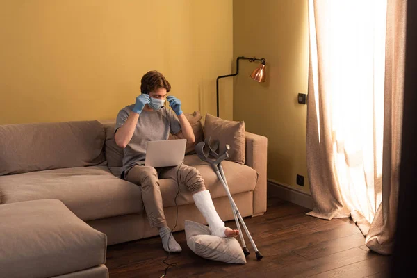 Freelancer em máscara médica com perna quebrada ajustando fone de ouvido perto de laptop e muletas no sofá — Fotografia de Stock