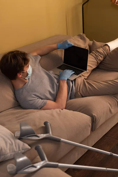 Mise au point sélective du pigiste dans un masque médical avec jambe cassée à l'aide d'un ordinateur portable près de béquilles sur le canapé — Photo de stock