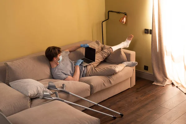 Freelancer em máscara médica e bandagem de gesso na perna usando laptop na sala de estar — Fotografia de Stock
