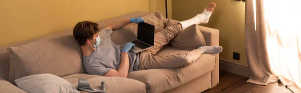 Panoramic concept of freelancer in medical mask with broken leg working on laptop at home — Stock Photo