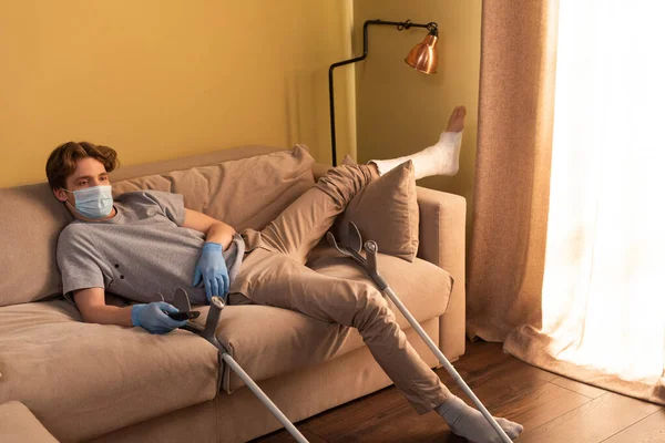 Disabled man in medical mask and plaster bandage on leg watching movie near crutches at home — Stock Photo