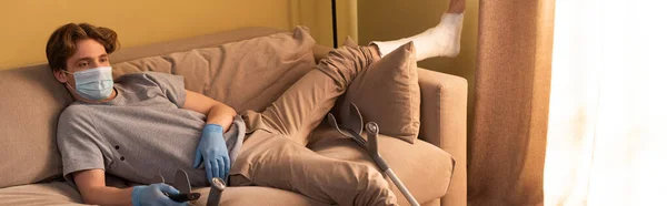 Horizontal crop of man in medical mask, latex gloves and plaster bandage on leg holding remote controller on couch — Stock Photo