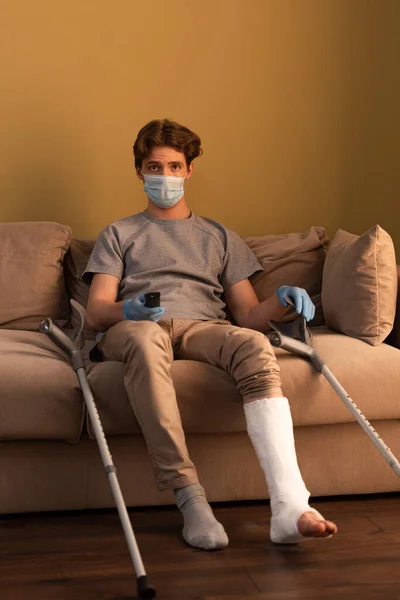 Hombre con máscara médica y vendaje de yeso en la pierna viendo la televisión mientras está sentado en el sofá en casa - foto de stock