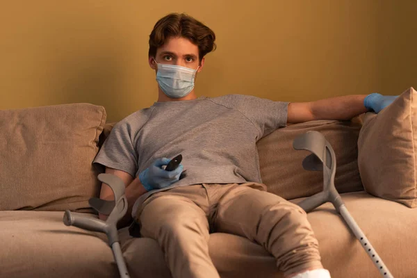 Foco seletivo do homem em máscara médica e luvas de látex segurando controlador remoto perto de muletas no sofá — Fotografia de Stock